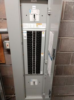 Electrical panel with closed door, mounted on a brick wall. Gray, with rows of breakers inside.