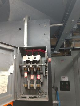 Electrical panel with open door, three red-labeled fuses, wires, conduit. Gray cabinet, industrial setting.