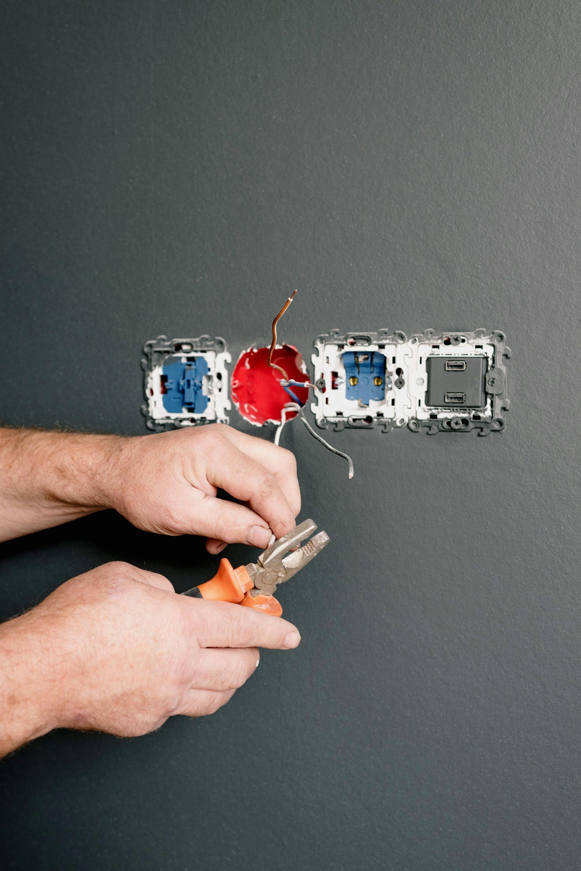 Electrician installing wiring in a wall outlet box with exposed blue, red, and gray electrical boxes.