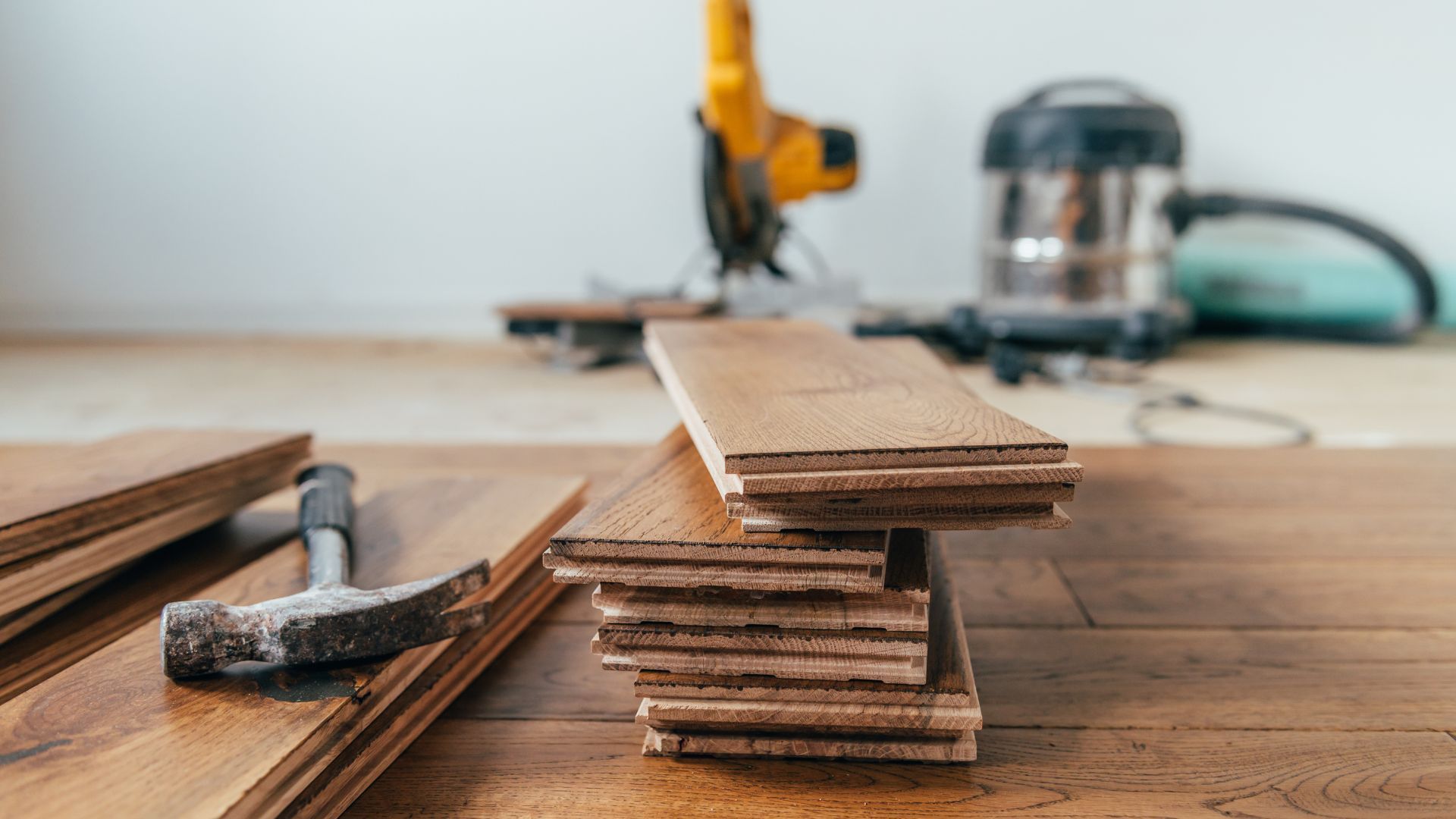 A hammer , a saw , and a vacuum cleaner are on a wooden table.