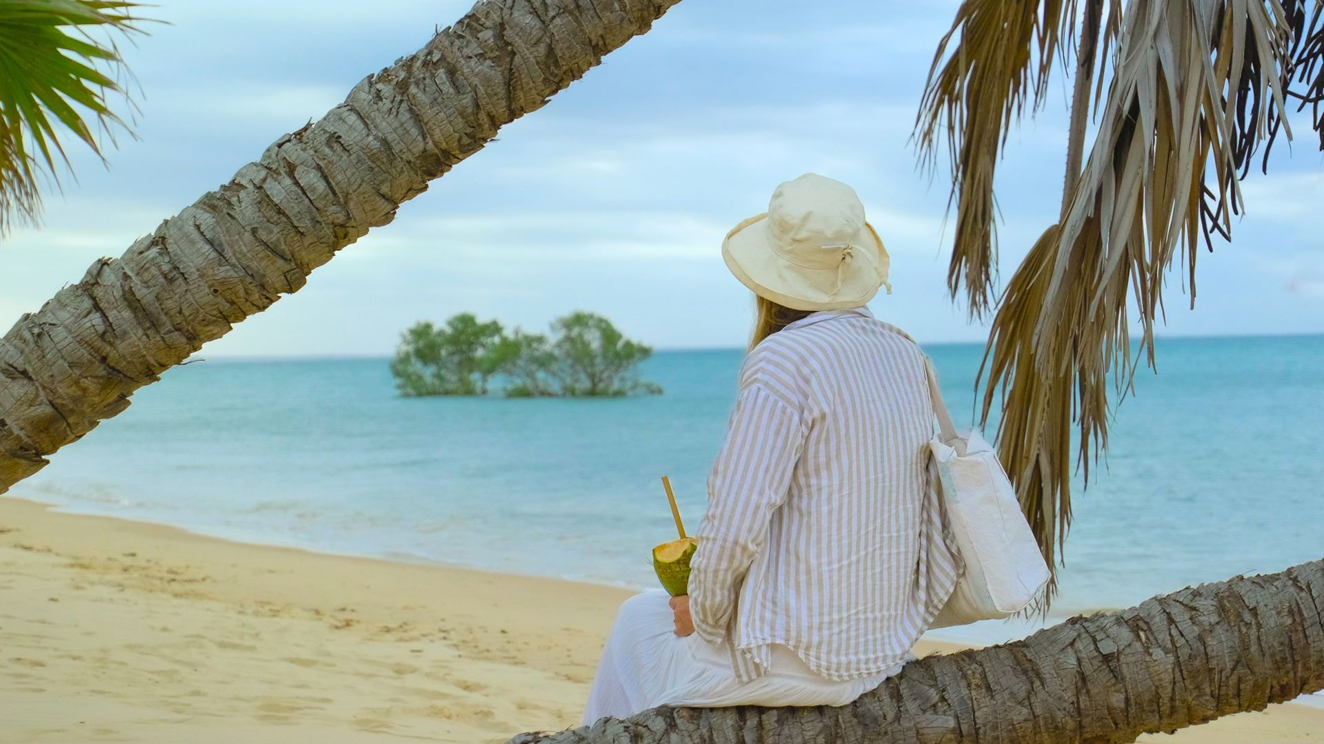 Person in a sunhat sits on a beach, facing the ocean with a small island in the distance.
