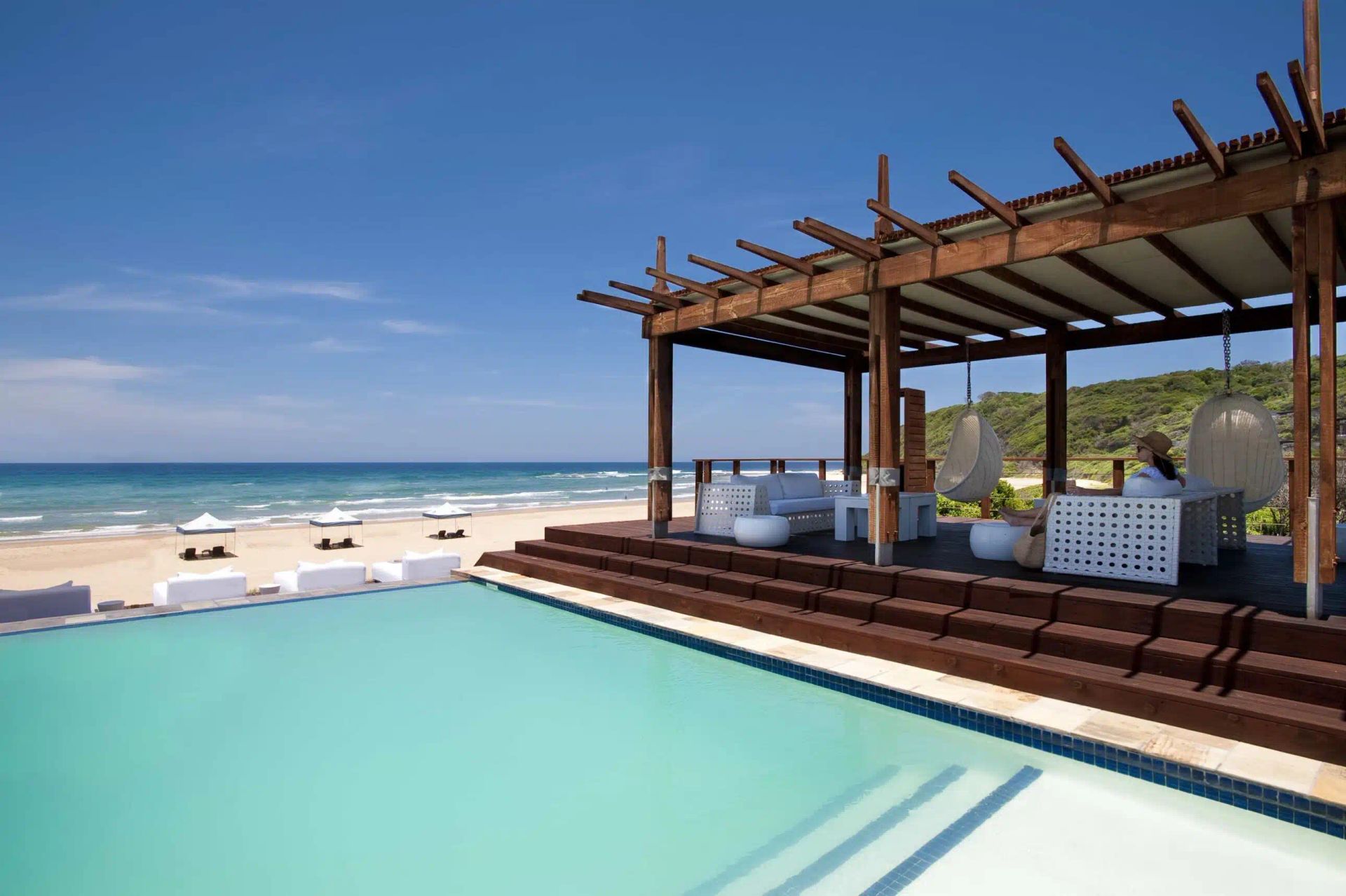 A wooden pergola with lounge seating overlooks a turquoise infinity pool and a sandy beach under a clear blue sky.