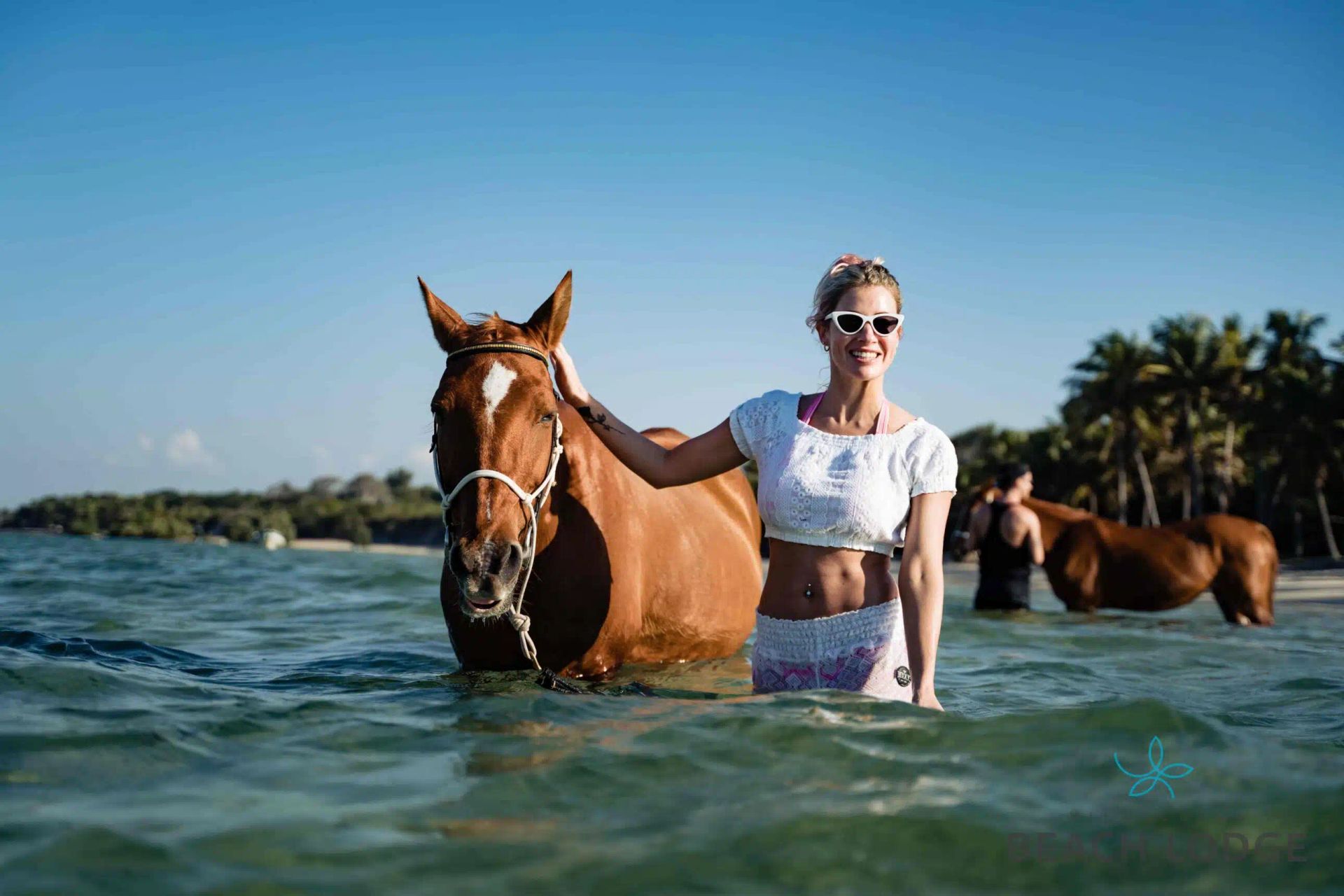 A person in a white top and sunglasses stands in the ocean with a brown horse, with another horse visible in the distance.