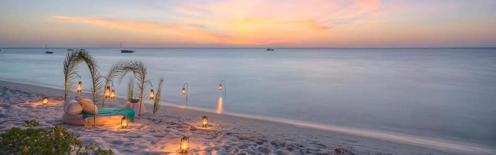 A romantic candlelit setup with arched floral decorations on a serene sandy beach at sunset overlooking the ocean.