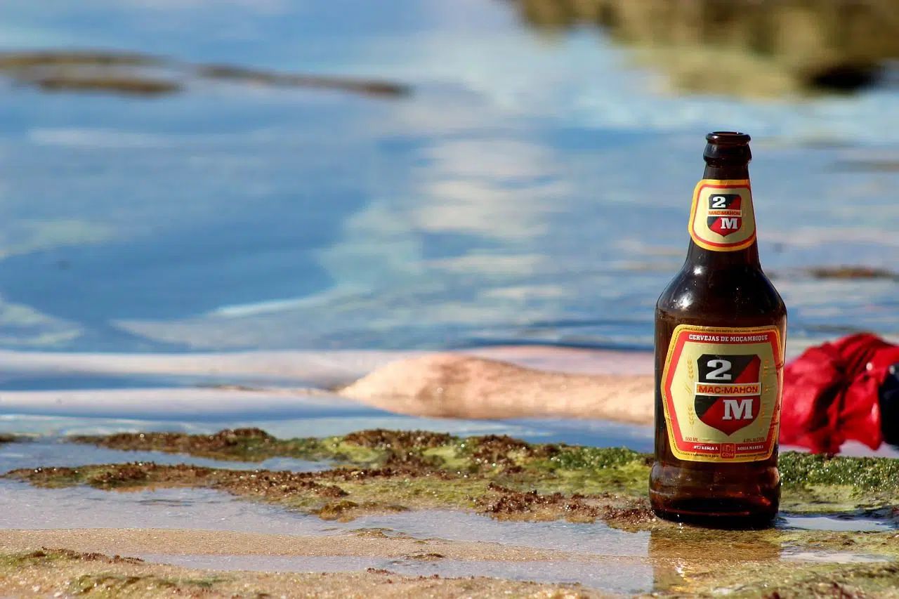 Beer bottle standing on a wet beach beside a red towel, with blue water in the background