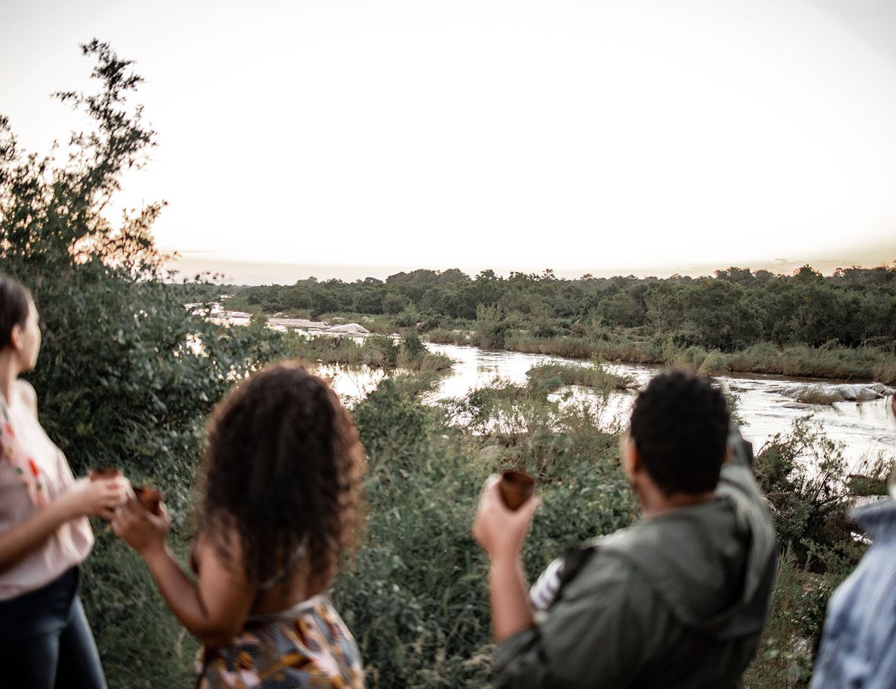 People holding drinks while overlooking a river winding through a lush, wooded landscape at dusk.