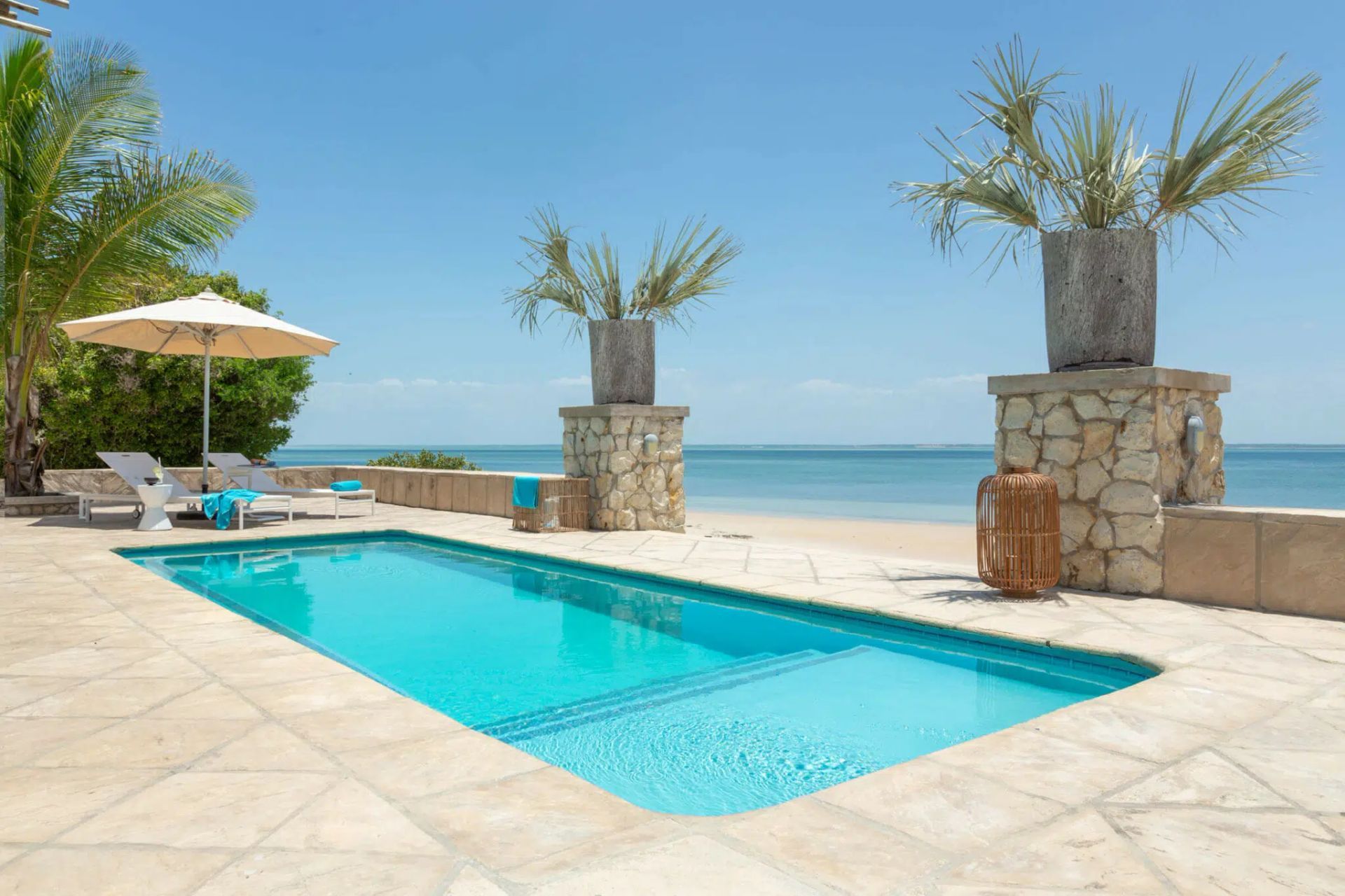 A bright blue rectangular swimming pool on a stone patio overlooking a calm ocean under a clear blue sky.