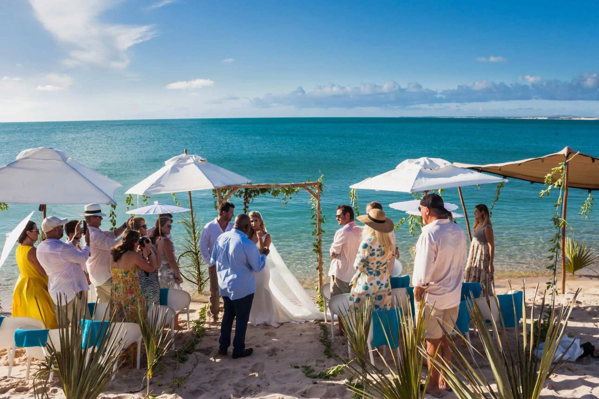 Beach wedding ceremony under white umbrellas beside turquoise ocean, with guests gathered around a floral arch.