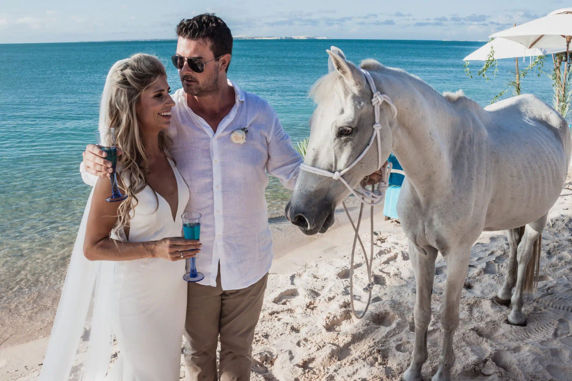 Couple standing on a beach beside a white horse with blue ocean in the background