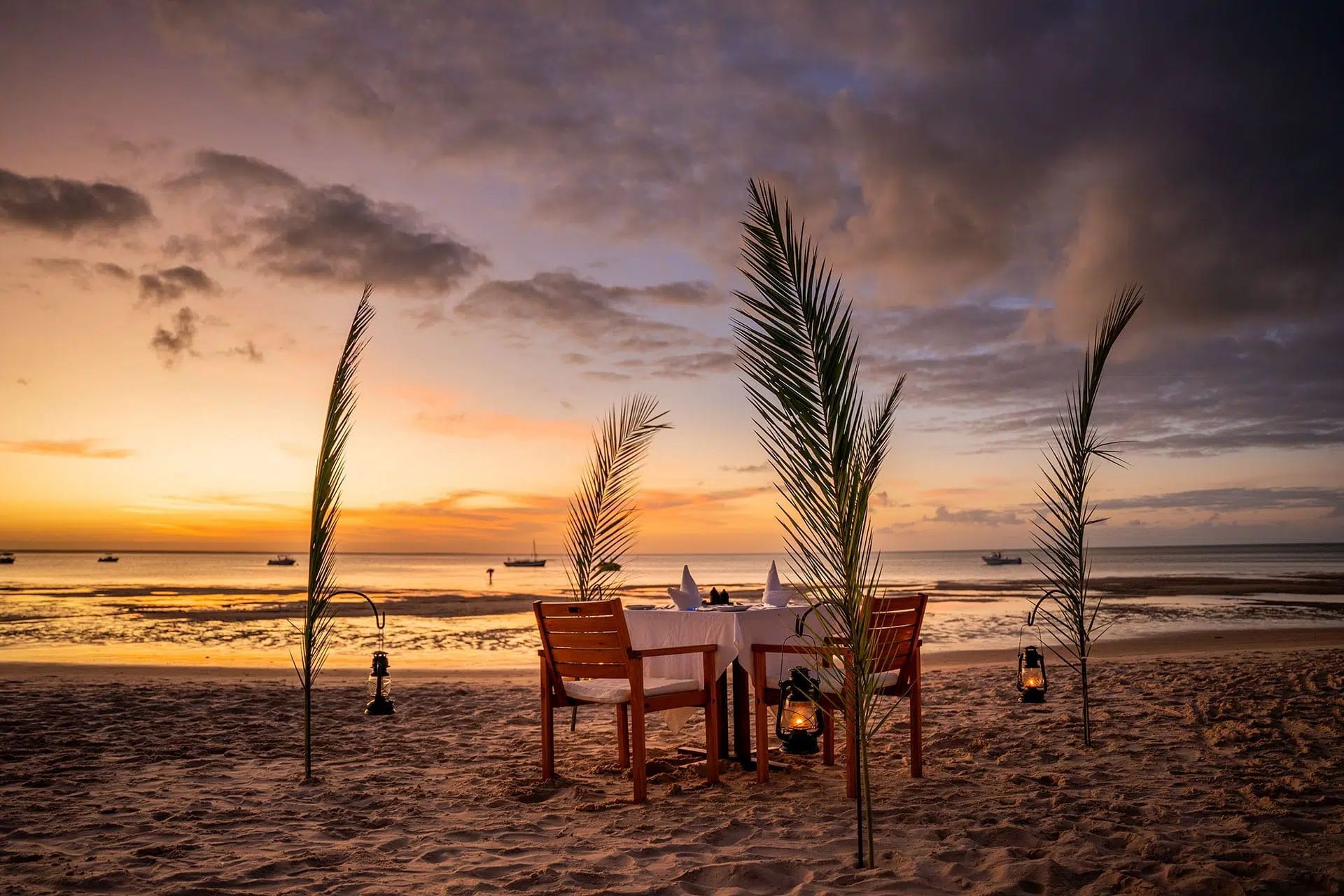 Beachside dining setup at sunset with a wooden table, chairs, lanterns, and tall grasses by the shore