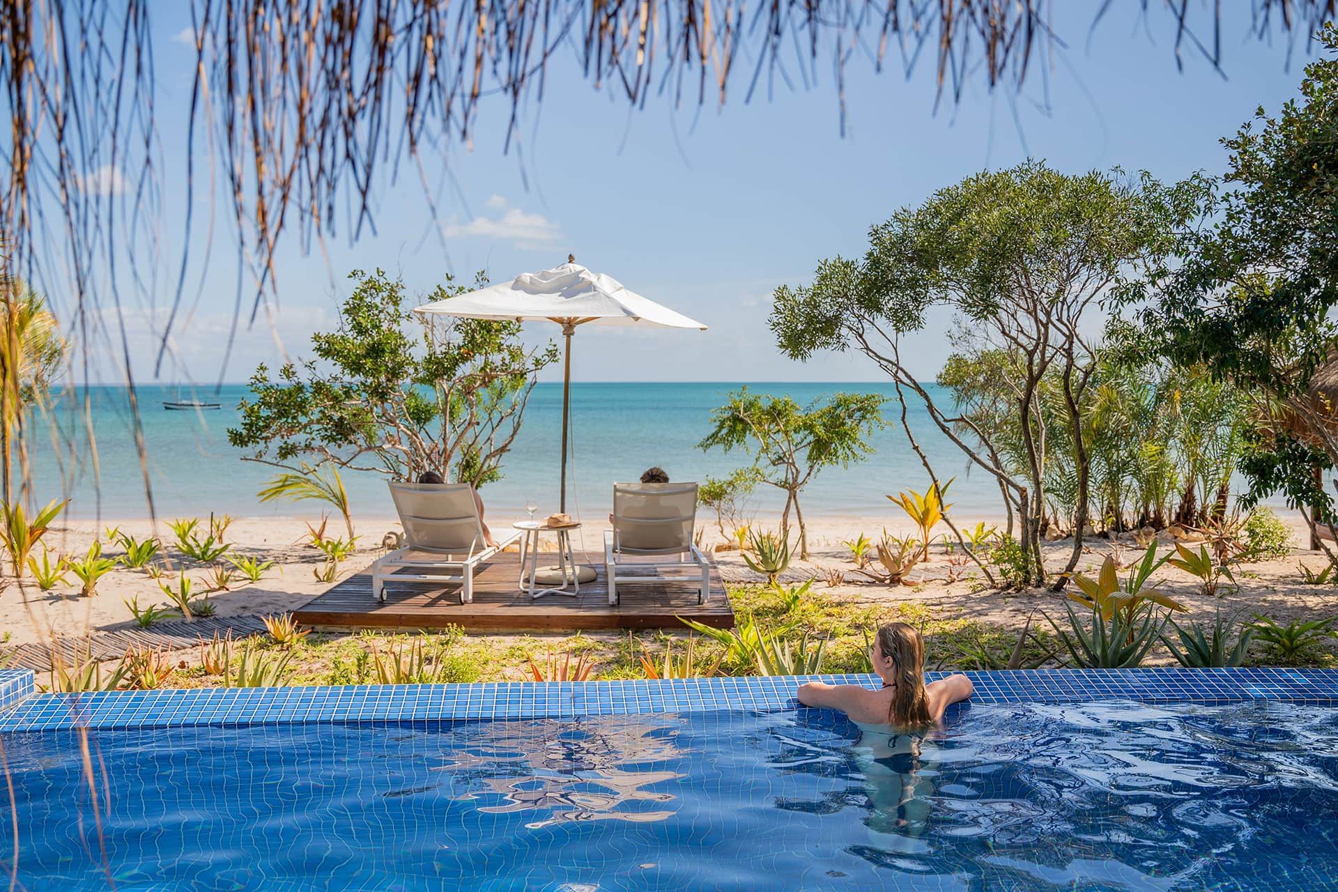 Infinity pool overlooking a tropical beach with white umbrellas and loungers under palm trees