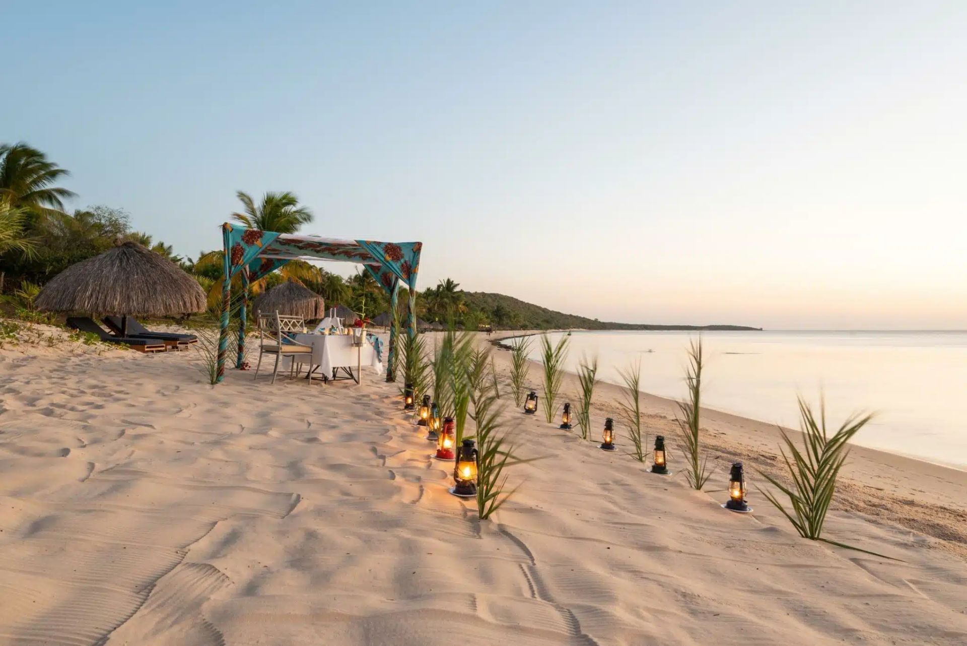 Beachside walkway with lit lanterns, palm trees, and a thatched hut at sunset by the sea