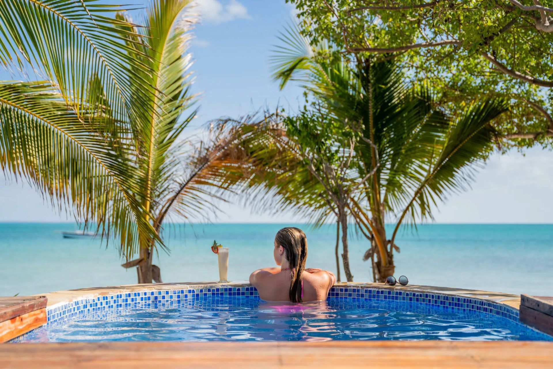 Person relaxing in a blue pool with palm trees and ocean view under a clear sky