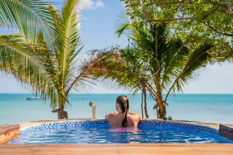 Person relaxing in a blue-tiled pool overlooking a tropical beach with palm trees and turquoise water