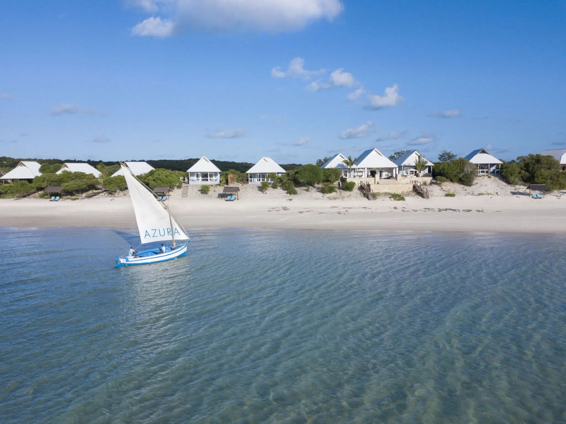 Sailboat gliding on calm blue water near a sandy beach with white houses and clear sky