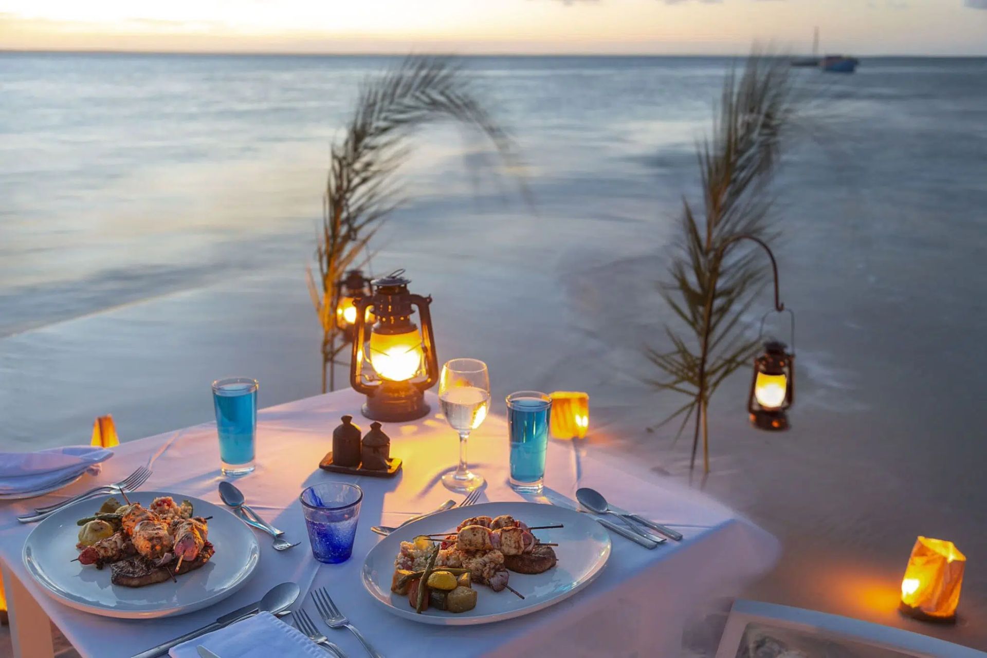 Romantic beach dinner at sunset with lit lanterns, blue drinks, and plates on a white tablecloth