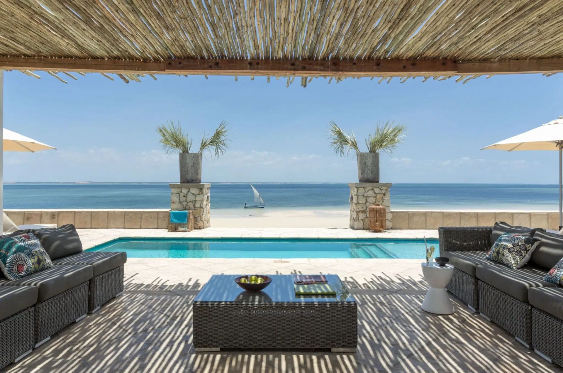 Shaded beach lounge with wicker sofas and table overlooking a pool and ocean, with sailboats in the distance