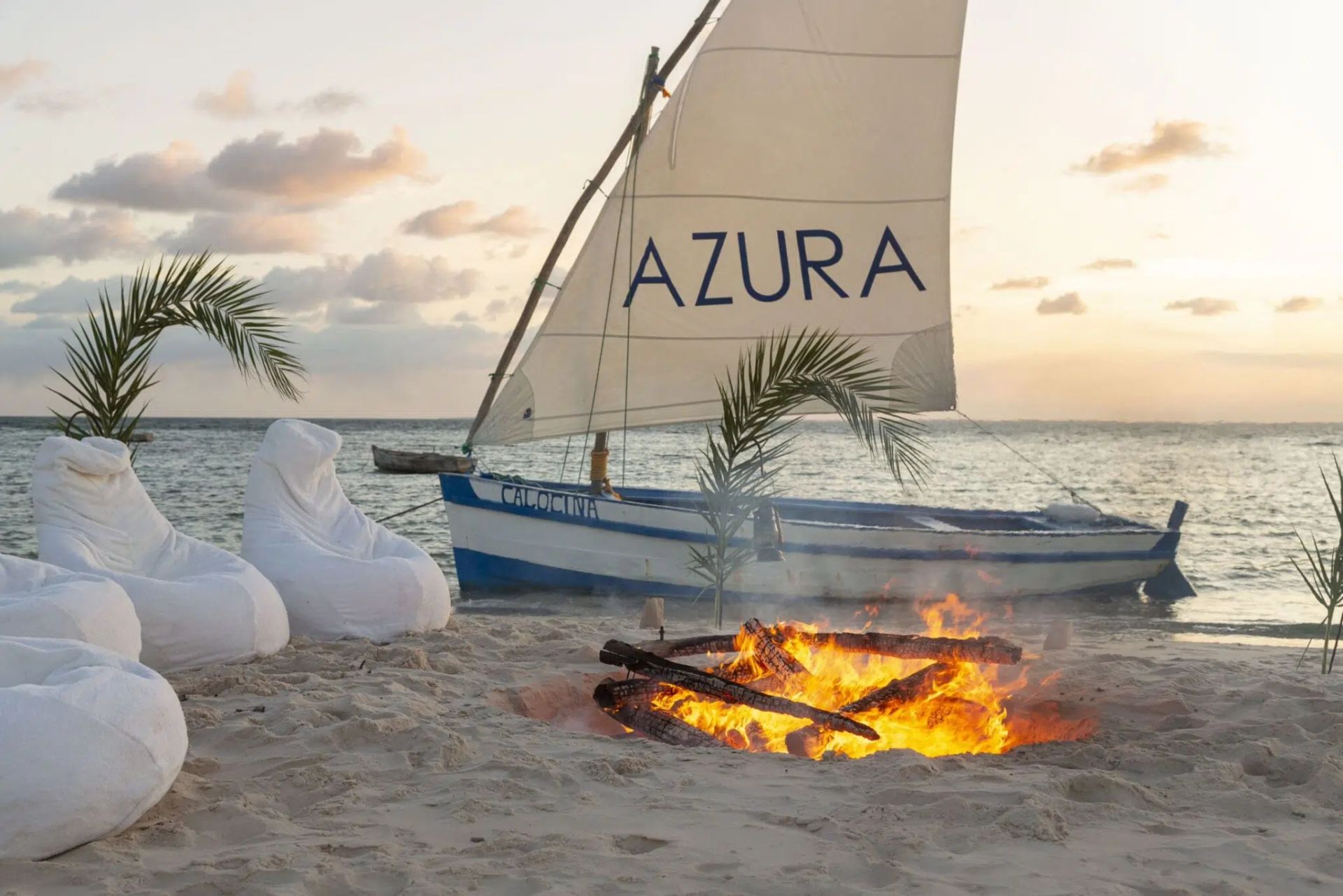Beach sunset with a sailboat named AZURA near a campfire and white bean bags on the sand