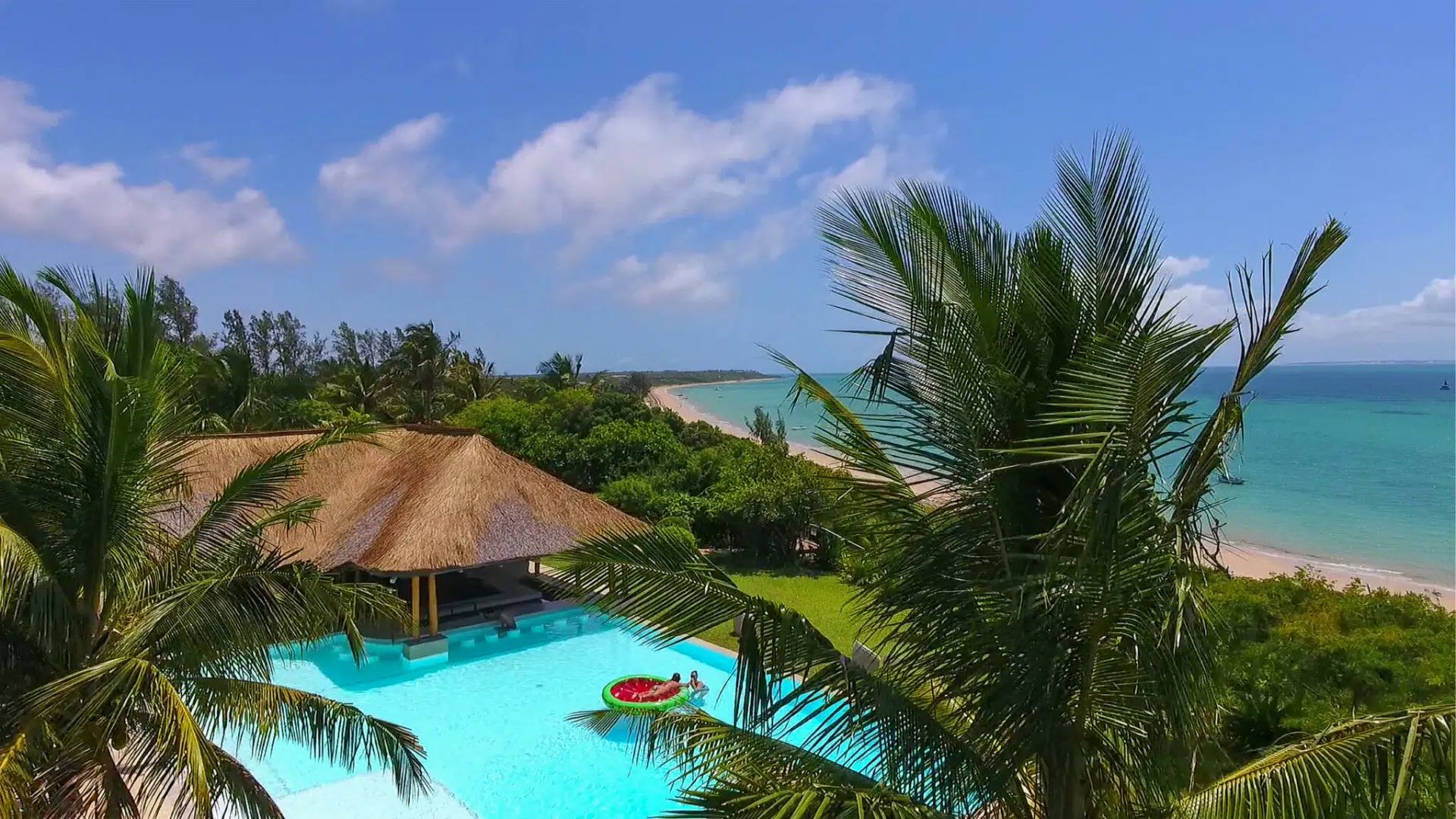 Tropical resort pool with thatched roof and palm trees overlooking a turquoise beach and sea