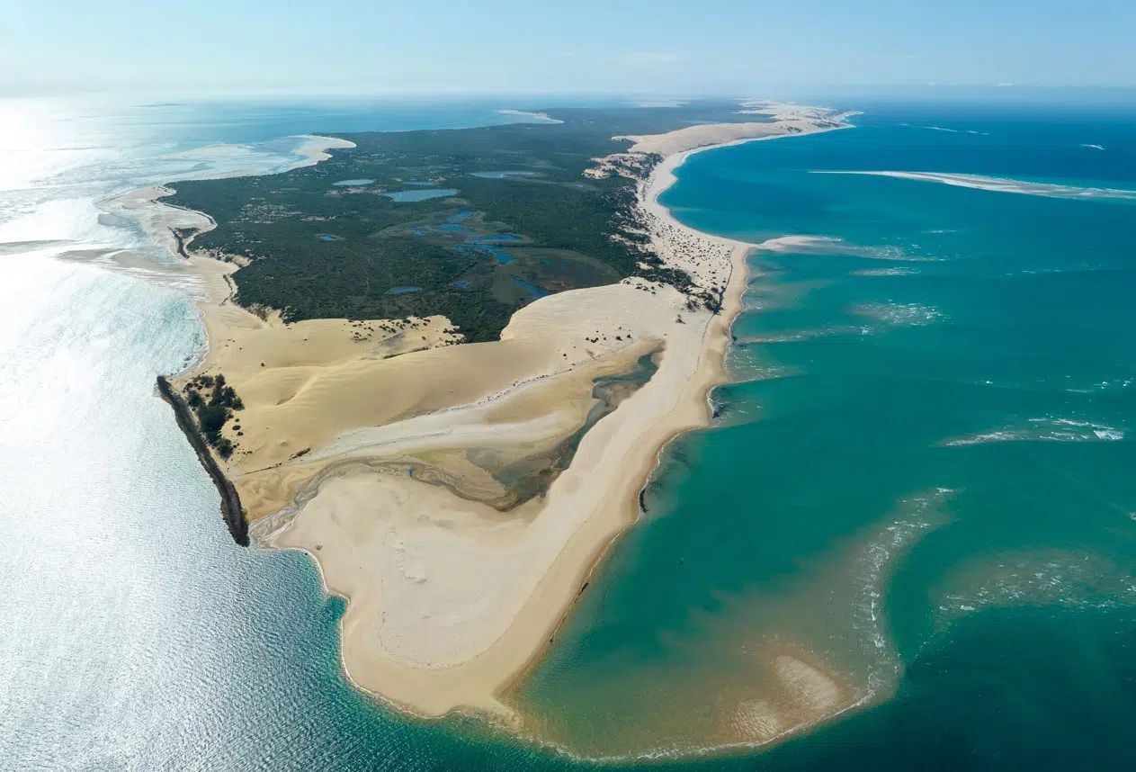 Aerial view of a sandy coastal peninsula between turquoise ocean and white surf, with dark green land inland