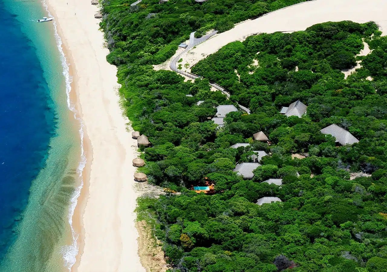 Aerial view of a tropical beach with turquoise water, white sand, and dense green trees with thatched huts.