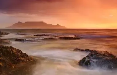 Rocky shoreline at sunset with waves, mist, and a distant island glowing in warm orange light