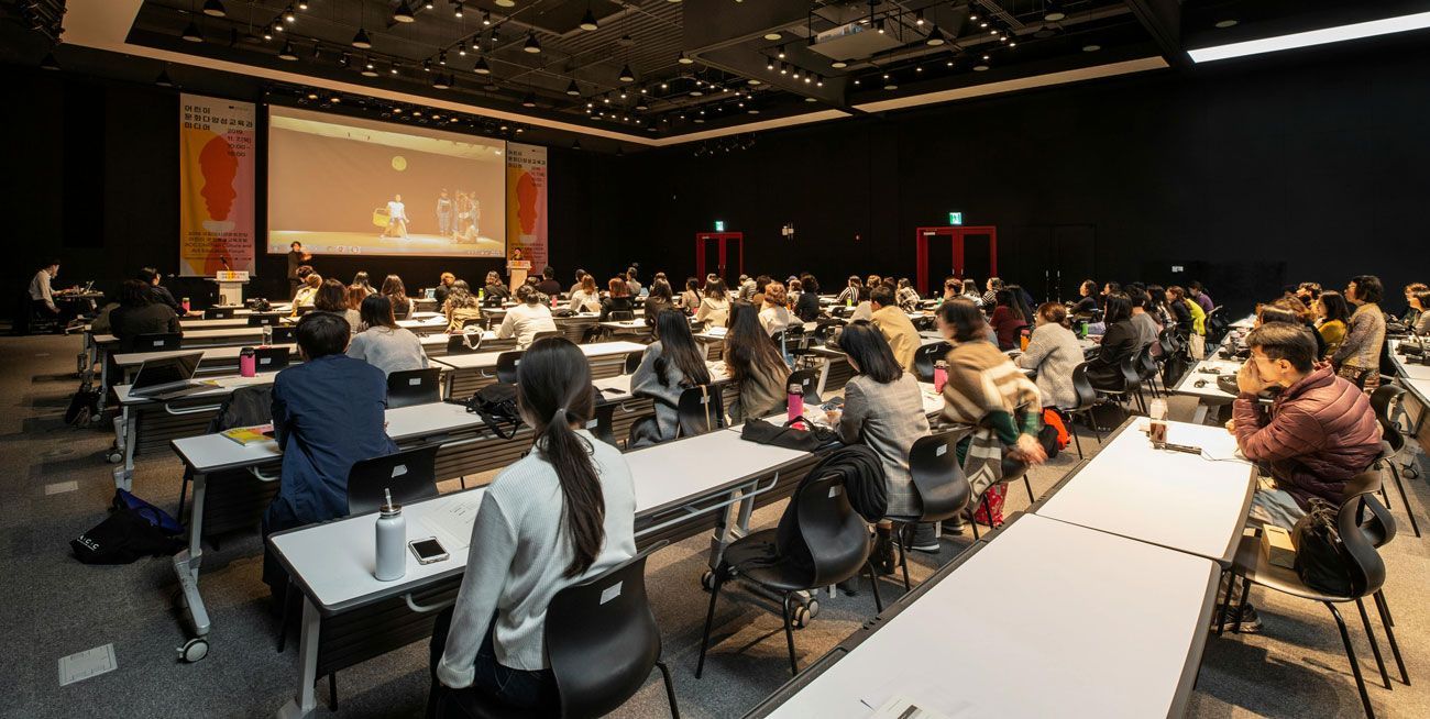 Audience seated in a large conference hall watching a presentation on a projection screen