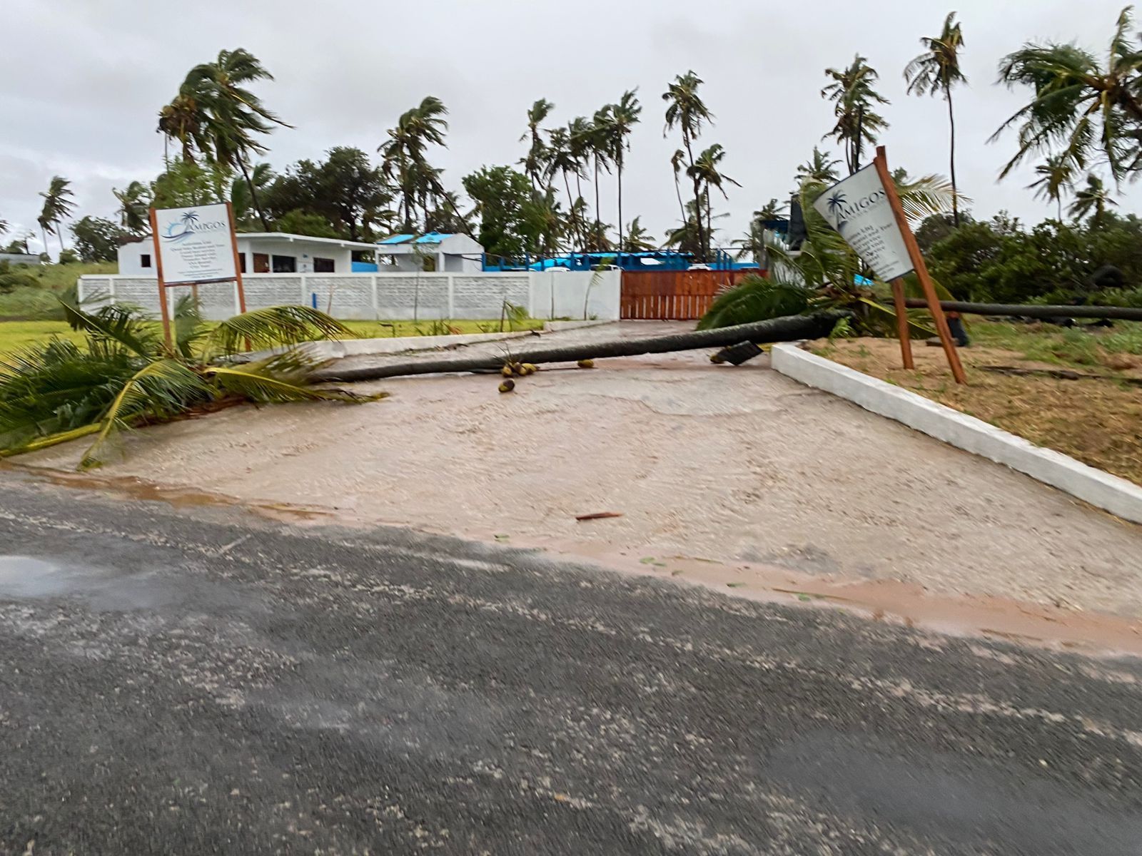 Flooded entrance road blocked by a fallen gate and leaning signposts near a coastal building.
