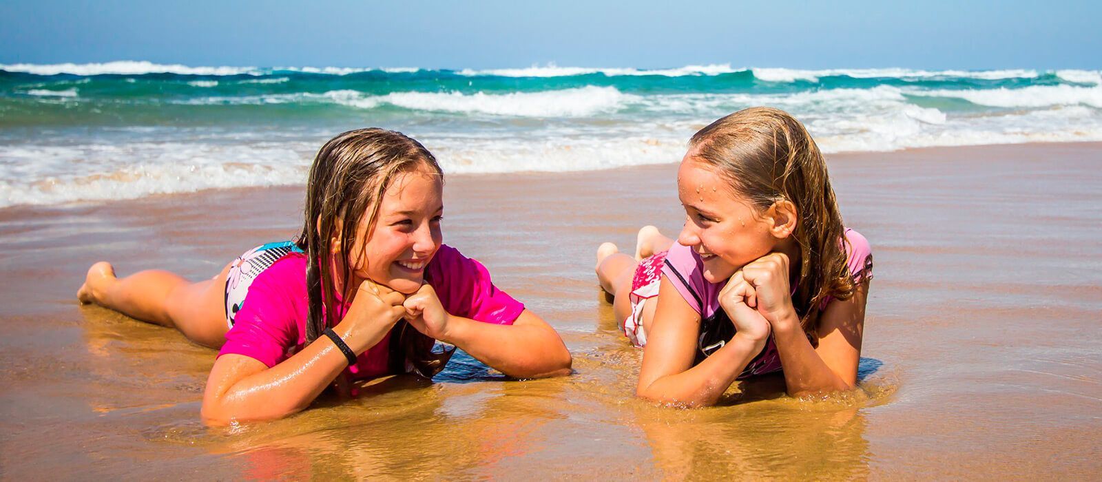 Two children in pink tops lying on wet sand, smiling at each other by the ocean.