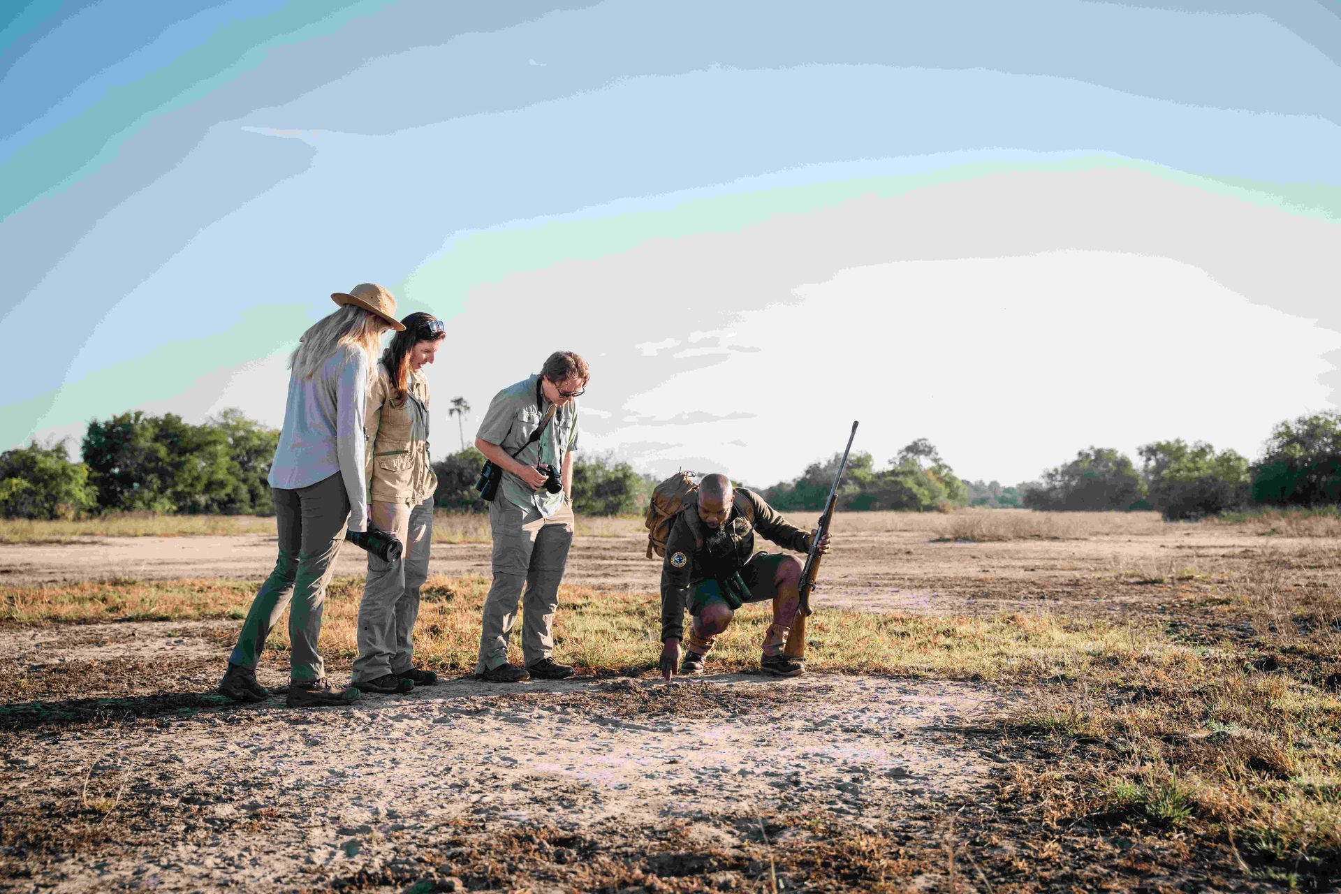 Four people outdoors examining a camera on a sunny, open field with dry grass.
