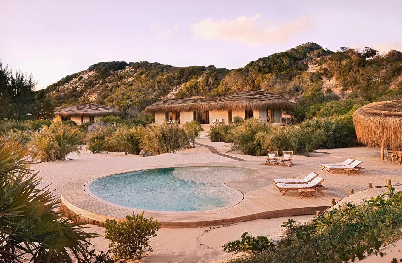 Resort courtyard with a round pool, lounge chairs, and stone buildings against a hillside at sunset