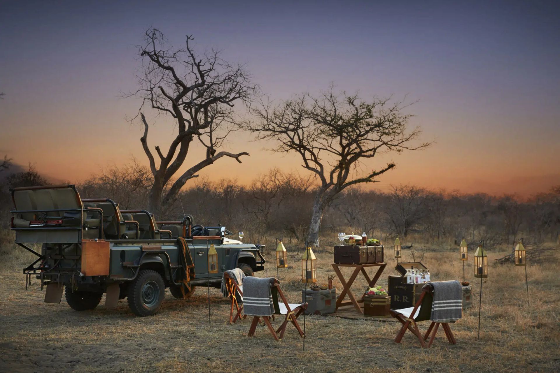 Safari camp with safari vehicles and tables under bare trees at sunset in a dry landscape