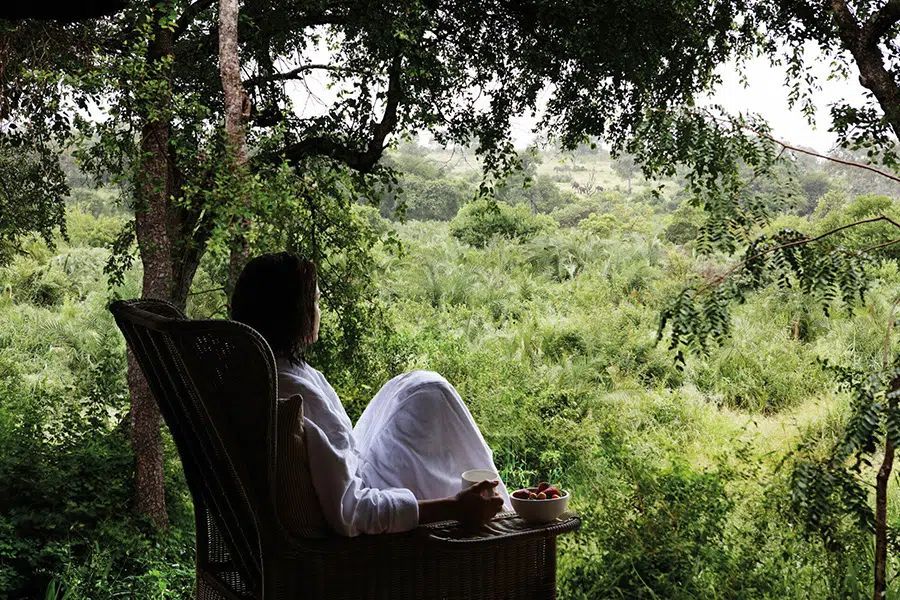 Person in a white outfit relaxing in a chair on a porch overlooking lush green countryside.