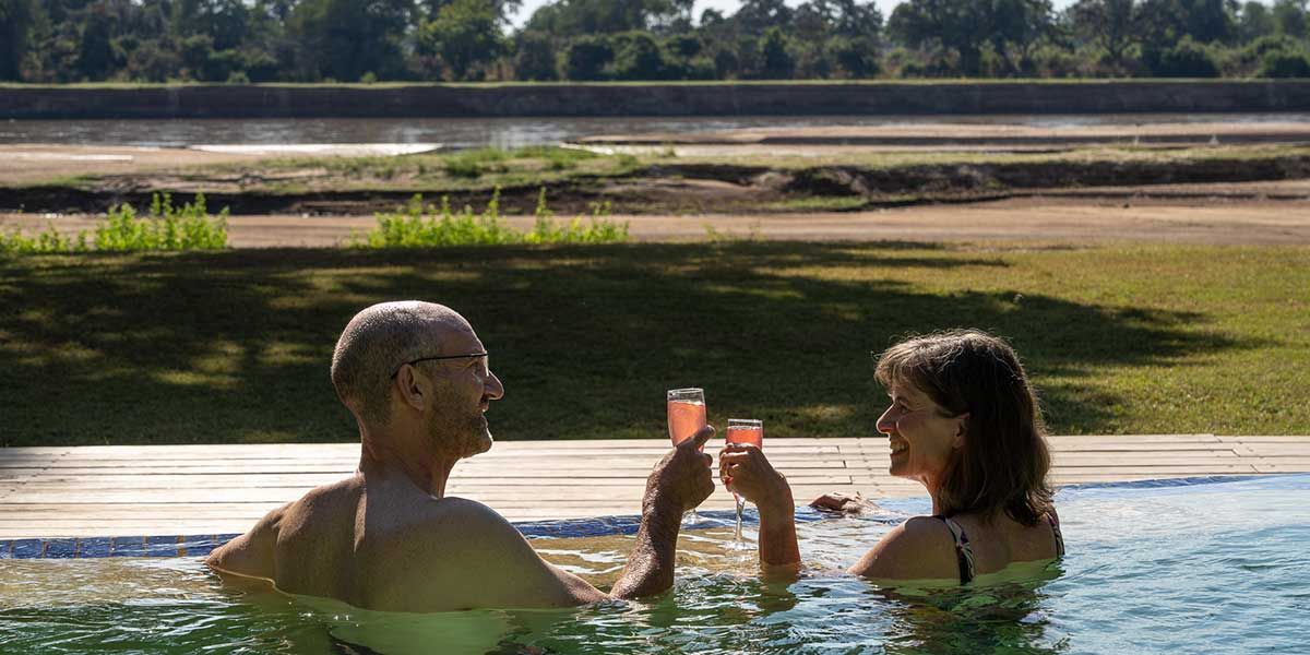 Two people in a pool clink drinks while overlooking a river or lake at sunset