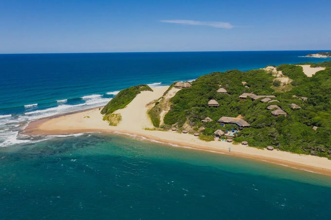 Aerial view of a tropical beach with turquoise water, white sand, and green hillside dotted with thatched huts