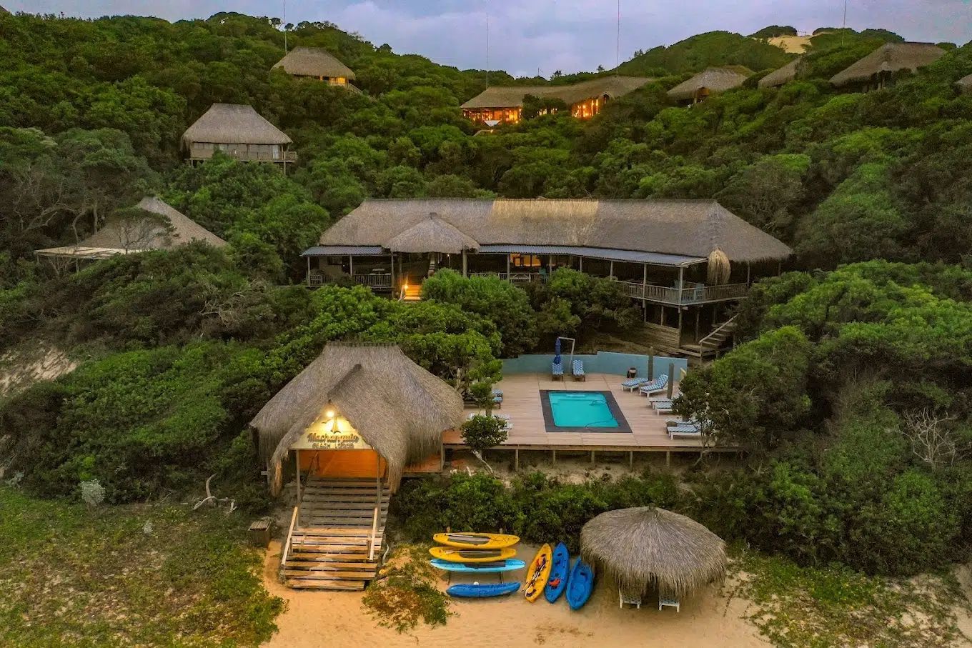 Thatched resort huts amid lush greenery, with a central pool and beach gear on sand