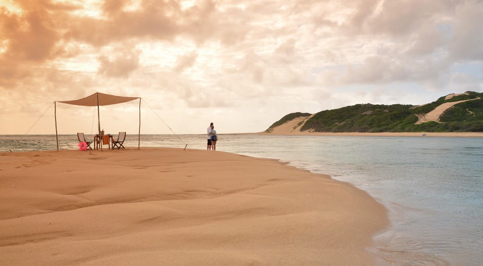 Sandy beach with a shaded umbrella, a few people, calm water, and a rocky coastline at sunset