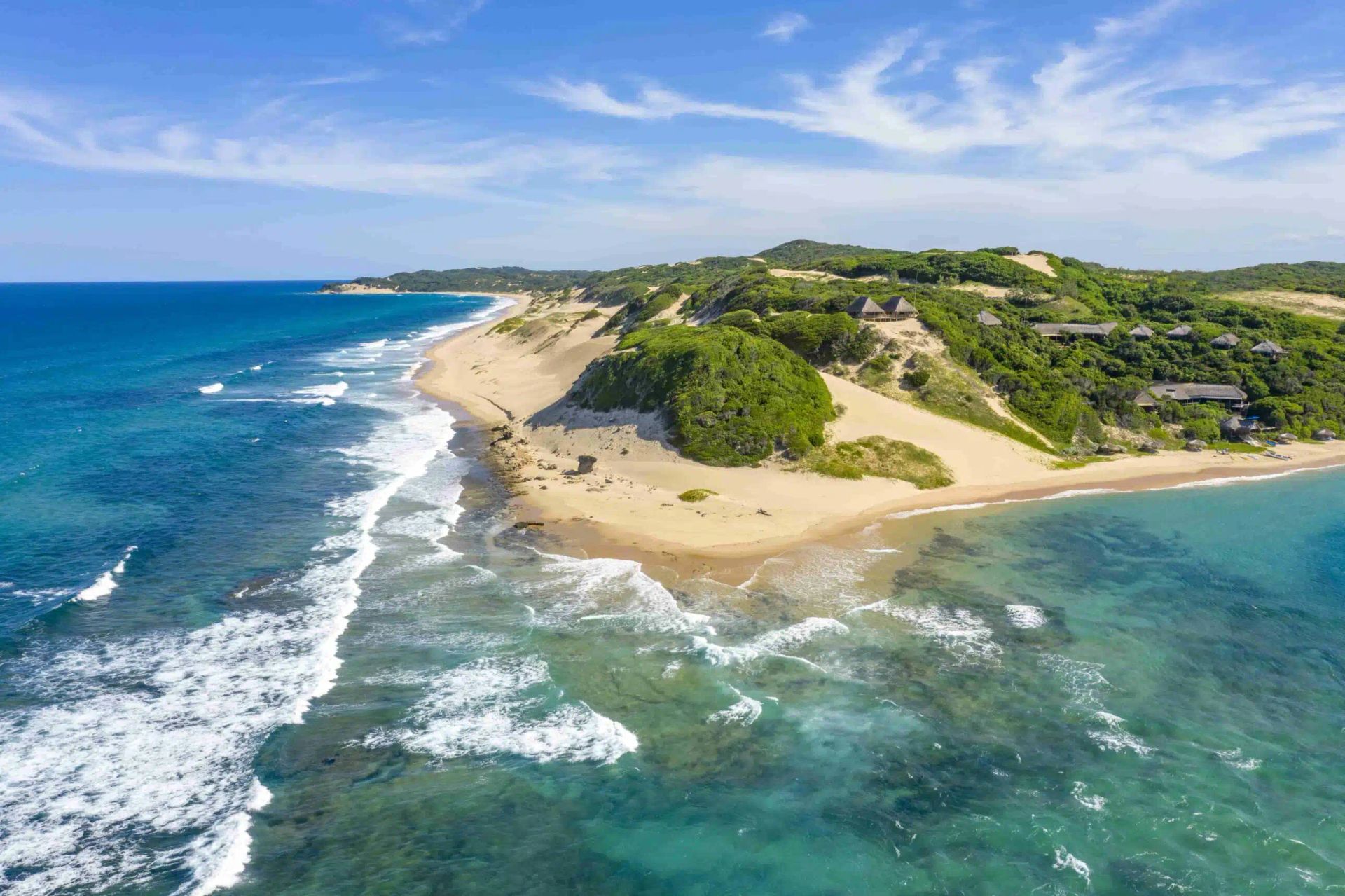 Aerial view of a sandy tropical peninsula with turquoise water, white surf, and green hillside vegetation