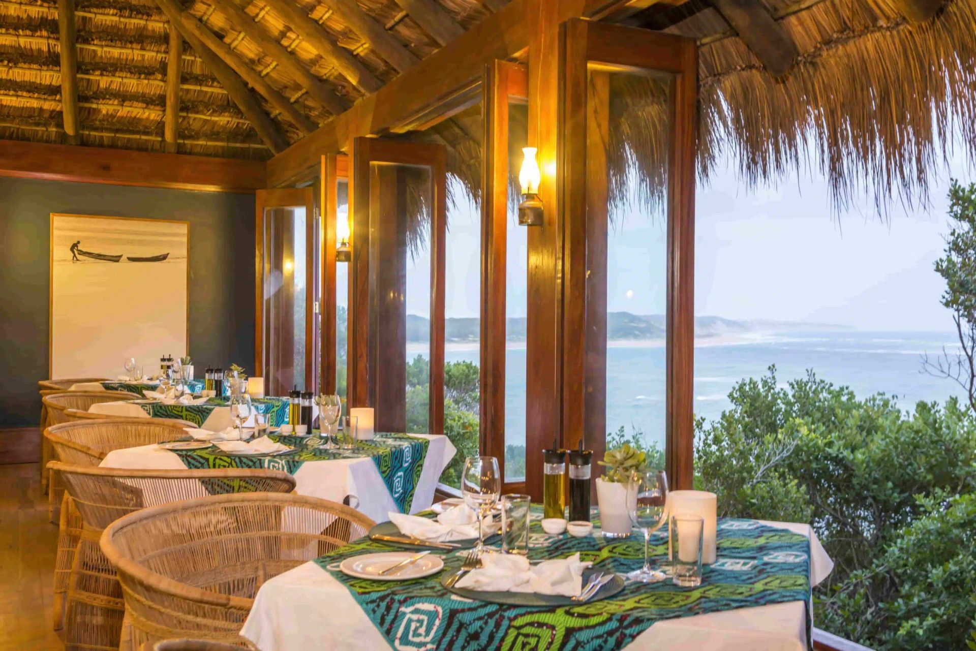 Open-air seaside dining area with wicker tables set for a meal, overlooking the ocean at sunset