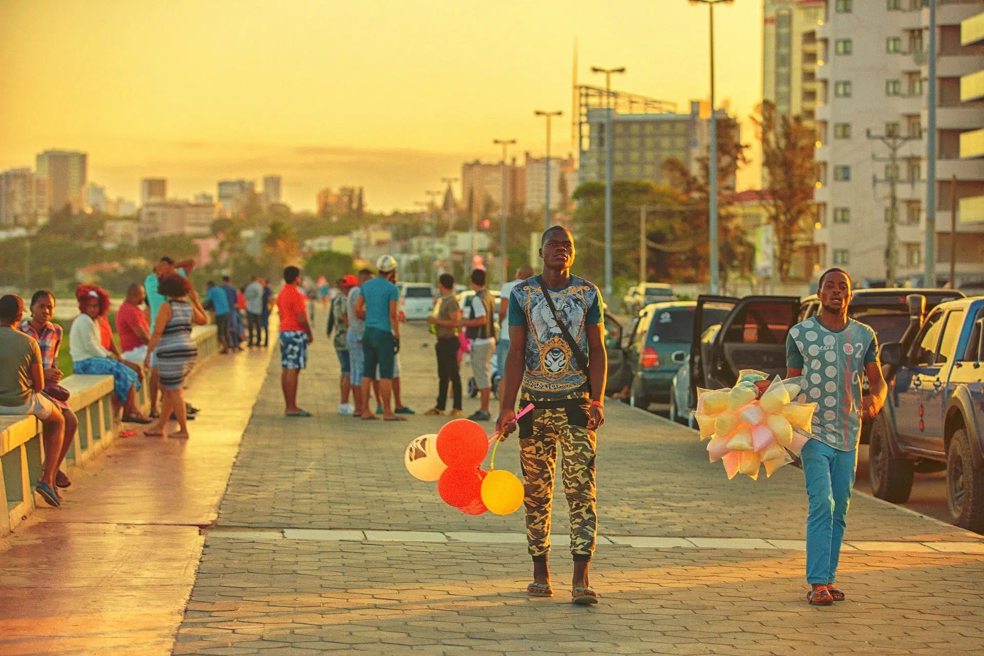 Two people walk along a sunset-lit waterfront promenade, one carrying colorful balloons.