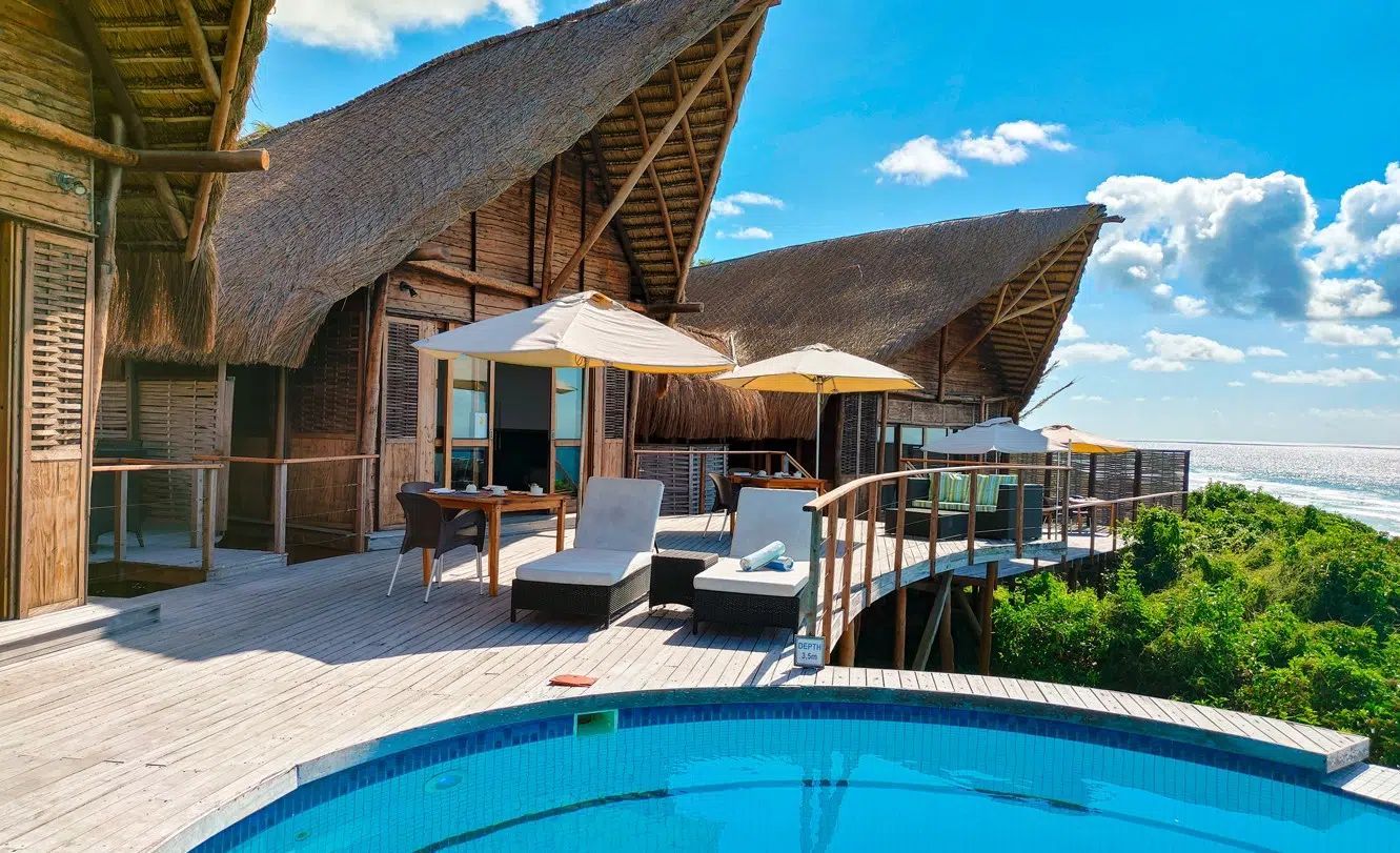 Poolside resort deck with lounge chairs, umbrellas, and thatched-roof buildings overlooking greenery and blue sky