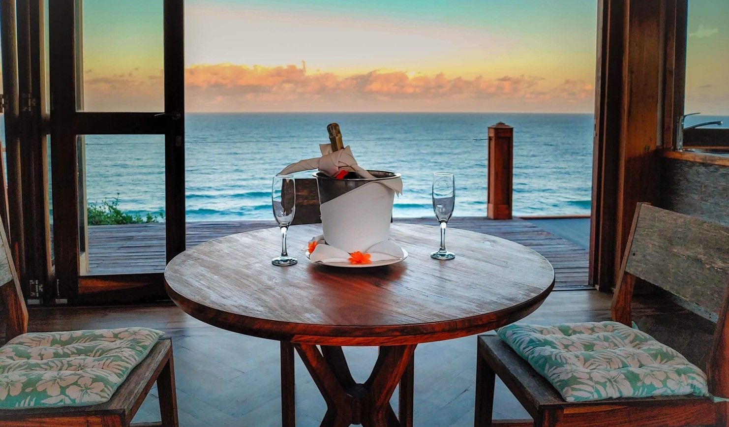 A seaside table with a bucket and bottle, framed by chairs, overlooking the ocean at sunset.