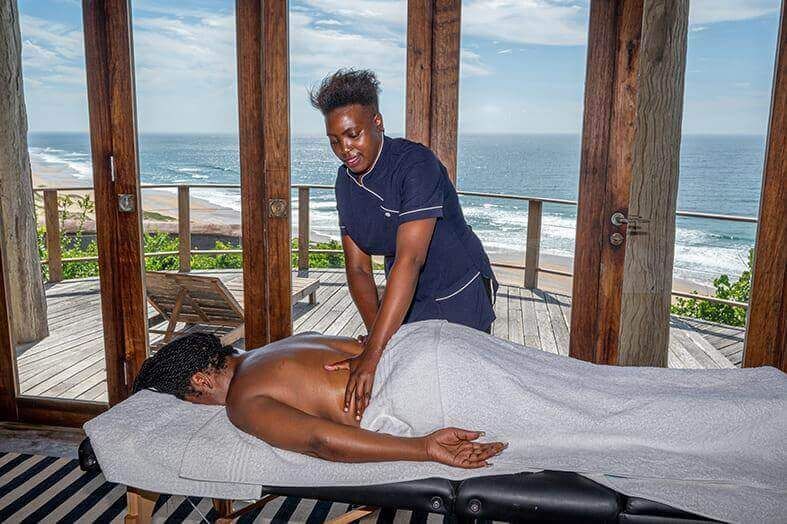 A massage therapist performs a treatment on a client lying on a massage table in a room overlooking the ocean.