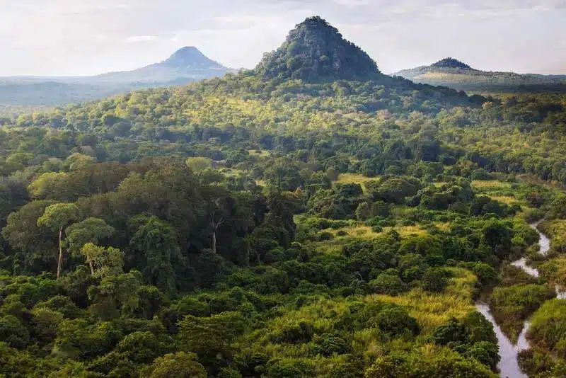 A vast, lush green landscape with three prominent, rounded mountain peaks rising above a dense tropical forest and river.