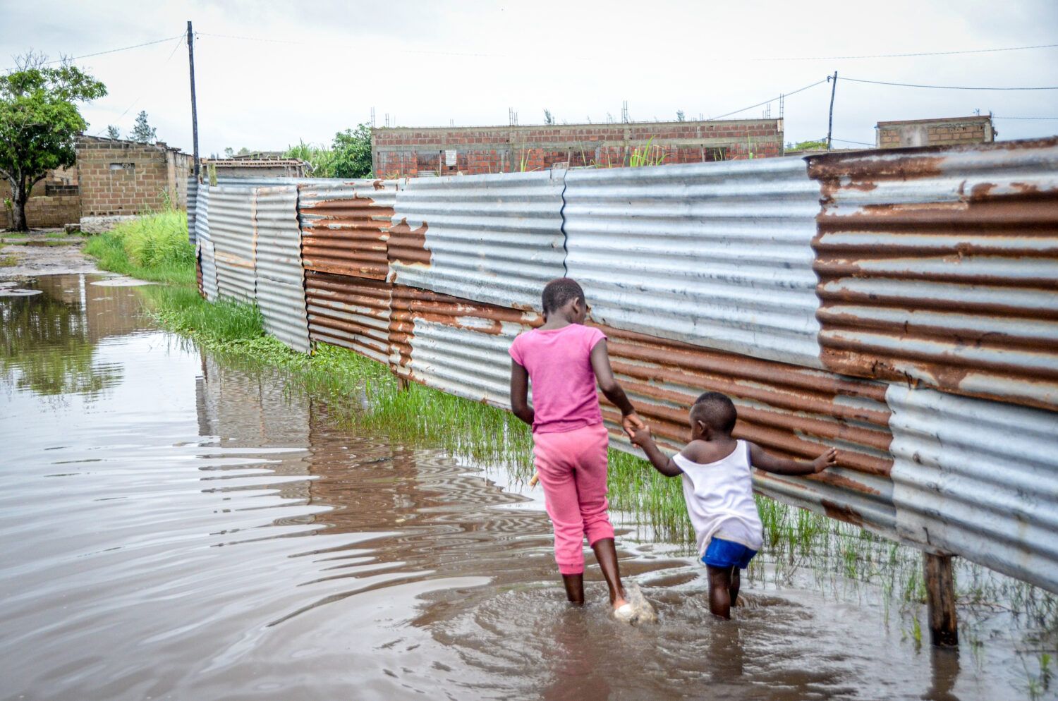 Two people wade through floodwater beside a corrugated metal fence.