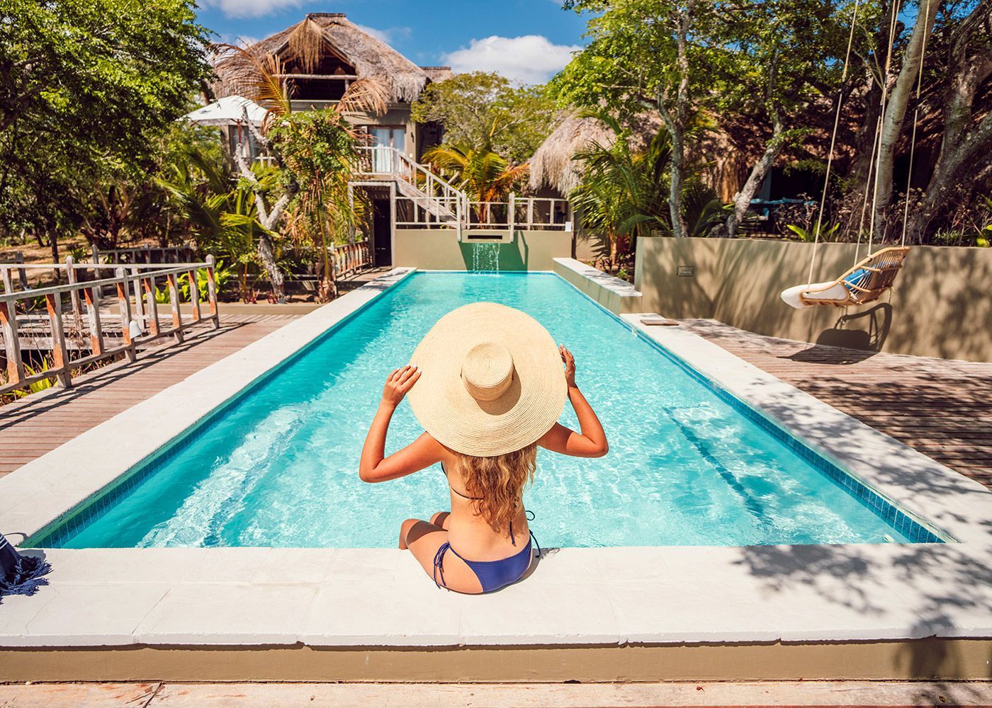 Person in straw hat sitting by a turquoise pool at a tropical resort, facing a thatched-roof building