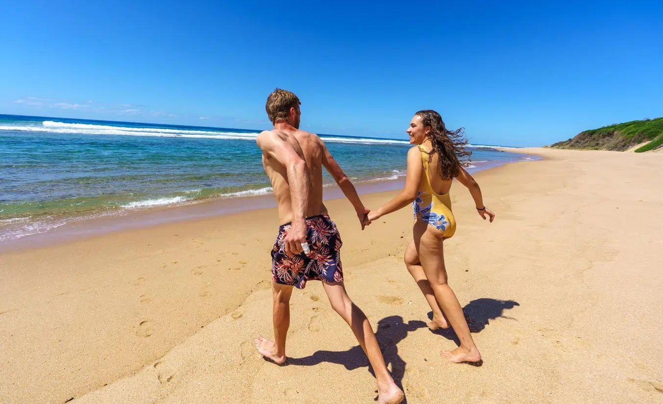 Two people holding hands and running along a sunny beach beside the ocean