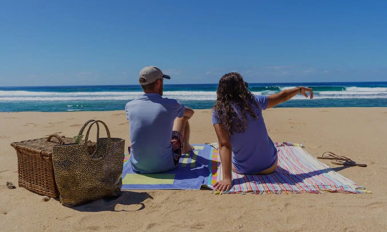 Two people sitting on a sandy beach overlooking blue water and a bright sky