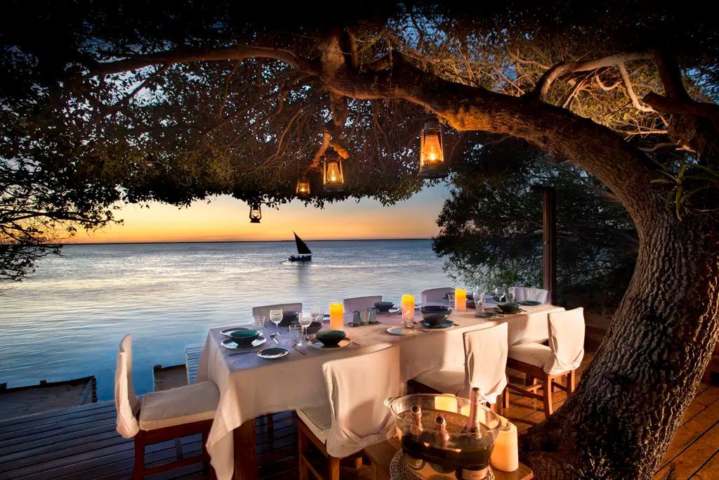Seaside dining table under a tree at sunset, with candles and a sailboat on calm water