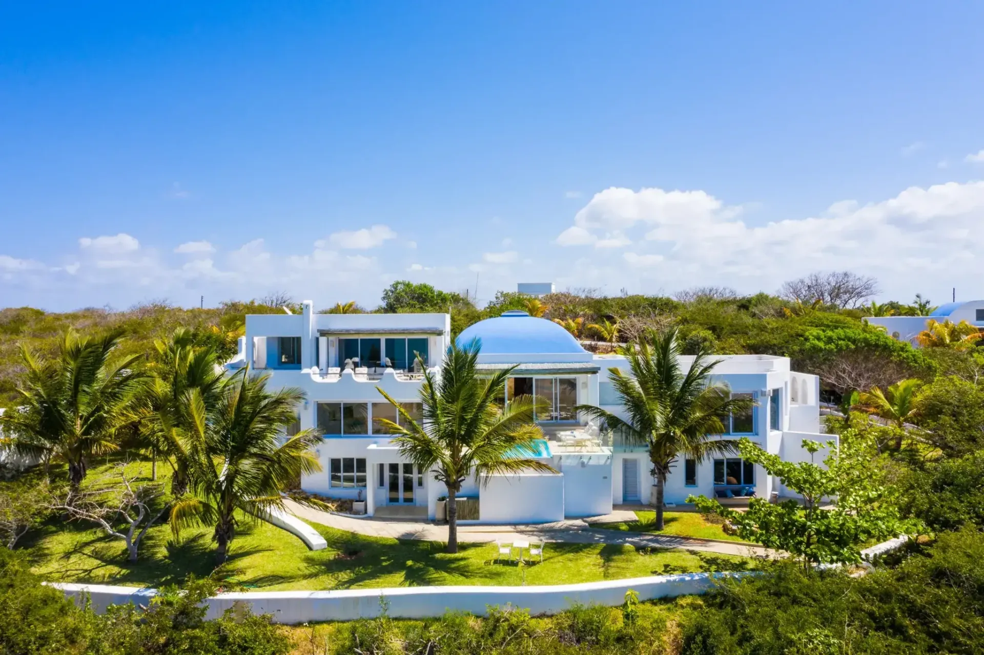 Modern white beachfront villa surrounded by lush tropical greenery under a clear blue sky